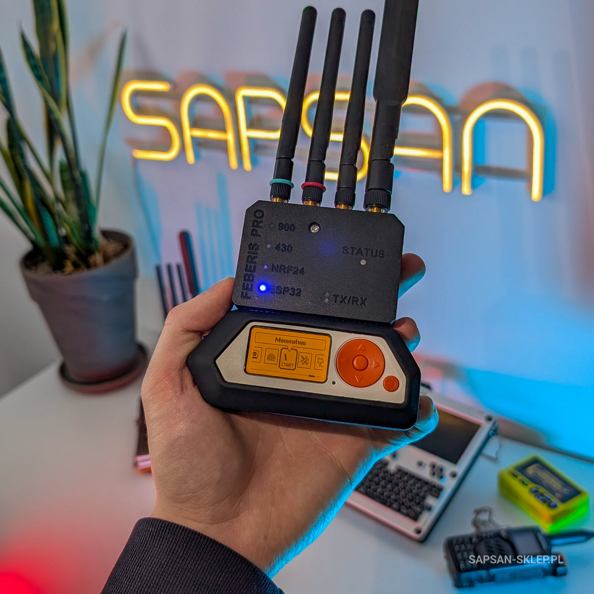 Flipper Zero z modułem Feberis Pro i czterema antenami w dłoni, w tle neon SAPSAN