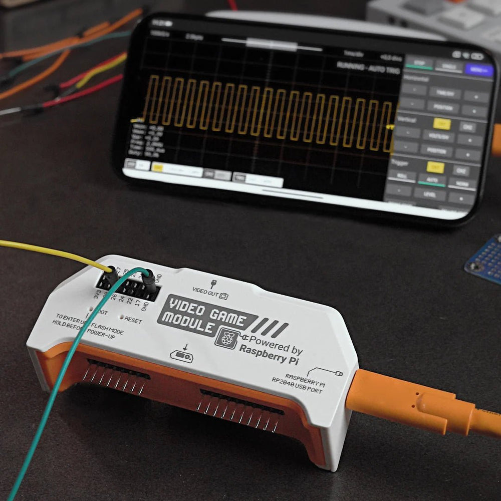 Игровой видеомодуль RP2040 на столе, подключённый оранжевым кабелем USB, на заднем плане смартфон с приложением осциллографа, показывающим прямоугольный сигнал