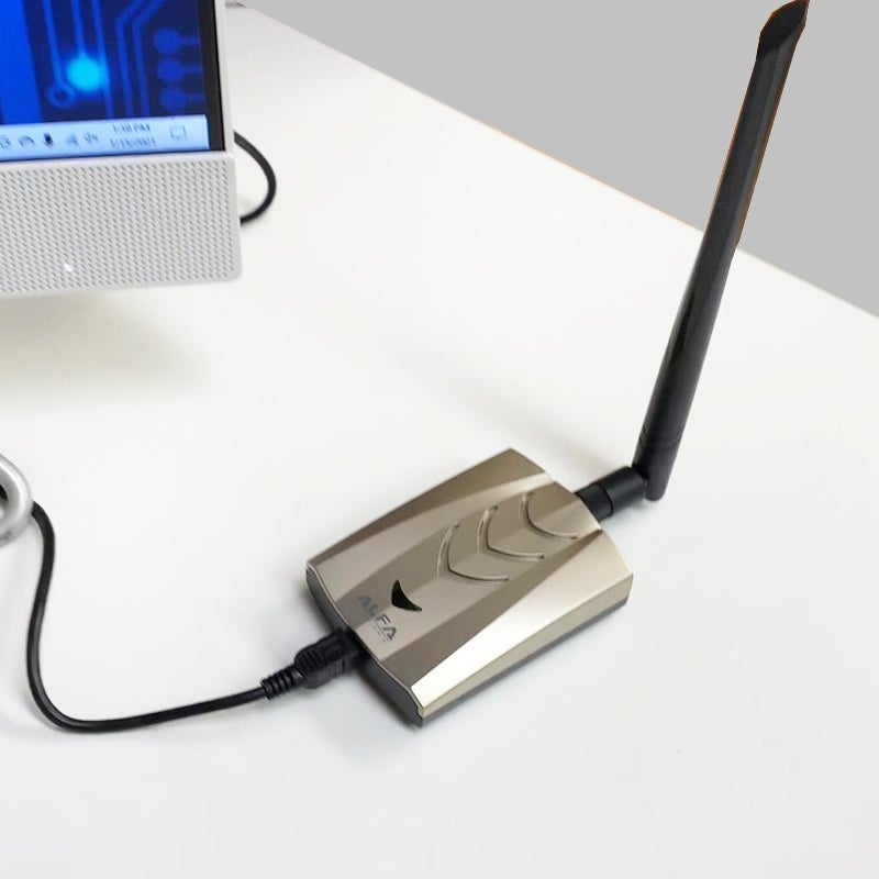 Silver and black Alfa AWUS036ACHM USB Wi-Fi adapter with antenna lying on a desk, connected by USB cable to a computer