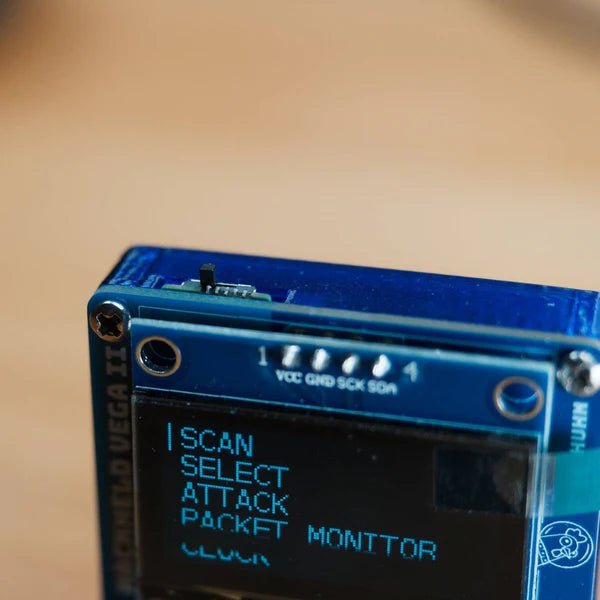 Close-up of the OLED screen of the HackHeld Vega II with a menu of SCAN, SELECT, ATTACK, PACKET MONITOR and CLOCK - a translucent blue case with an embossed HACKHELD VEGA II inscription, GPIO pin labels VCC GND SCK SDA numbered 1-4 and a Spacehuhn owl logo in the corner