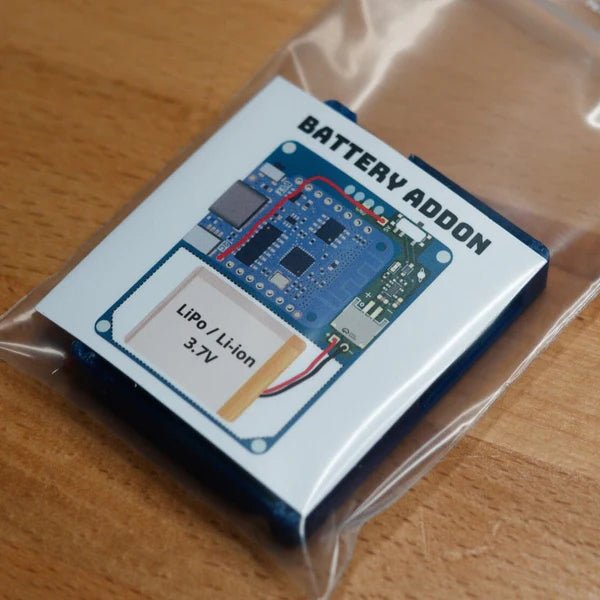 Battery Mod for the HackHeld Vega II in a clear anti-static bag on a wooden surface - the card shows the BATTERY ADDON title, a photo of the blue Vega II board with a LiPo battery wired up and a LiPo / Li-ion 3.7V battery graphic