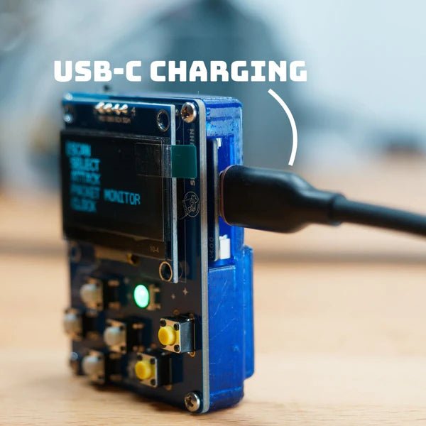 HackHeld Vega II with the Battery Mod being charged via a USB-C cable plugged in on the side on a wooden surface - a USB-C CHARGING caption with a hand-drawn arc, the OLED screen shows a menu with SCAN, SELECT, ATTACK, PACKET MONITOR and CLOCK, a glowing green LED and white and yellow buttons