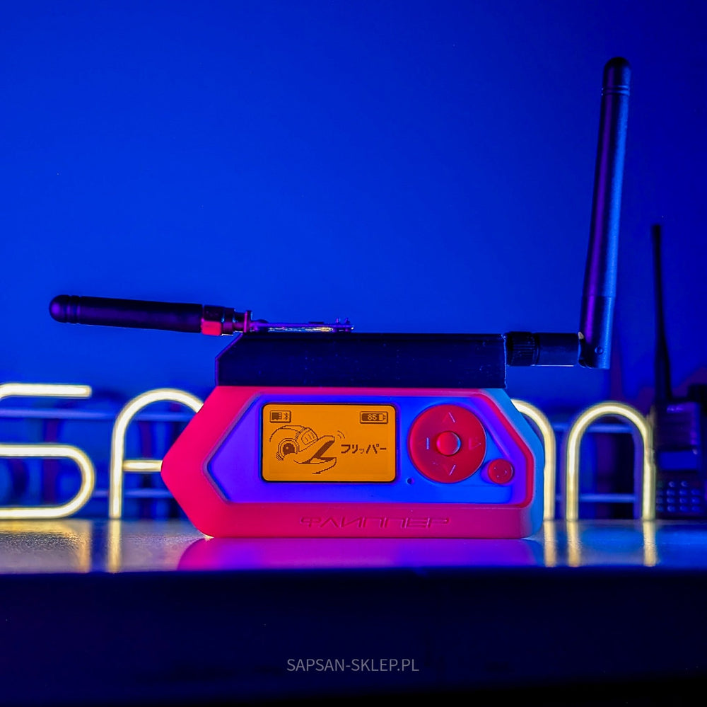A Flipper Zero in a pink case with the FEBERIS module and two antennas stands on a desk, the orange screen showing a dolphin with headphones and フリッパー text; behind it a yellow-glowing SAPSAN sign and a radio, SAPSAN-SKLEP.PL watermark