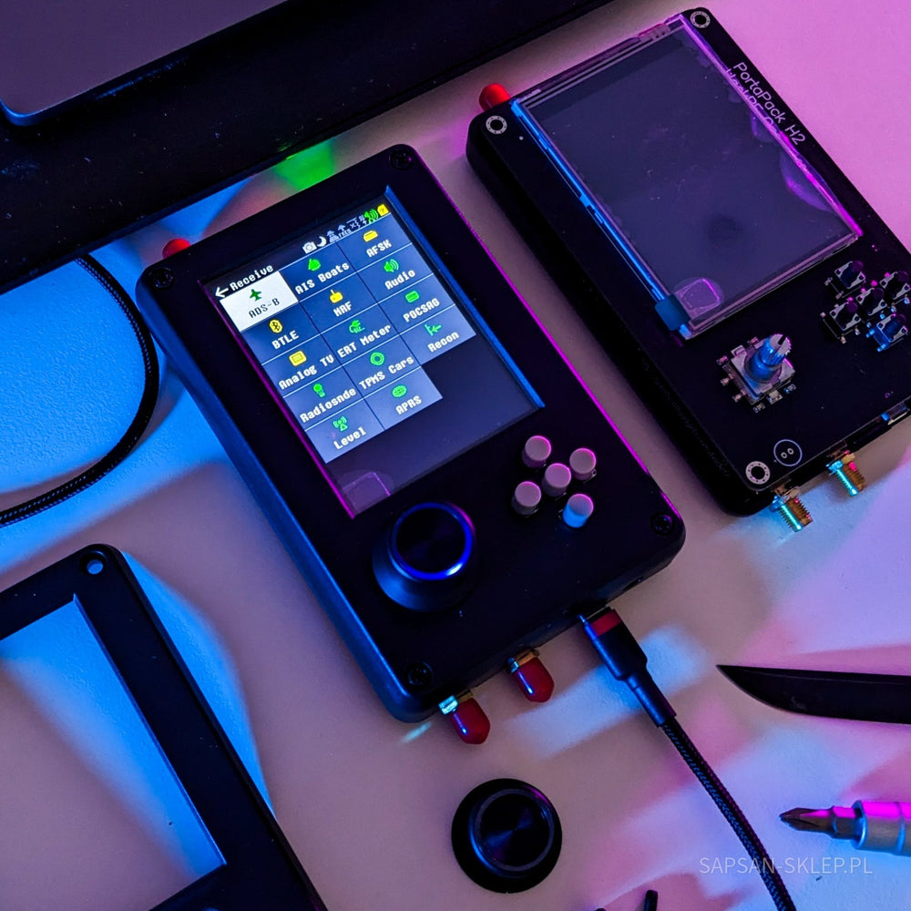 Černé zařízení HackRF One s nástavbou PortaPack H2 a dotykovou obrazovkou zobrazující menu příjmu (ADS-B, AIS, BTLE, Audio), v barevném osvětlení