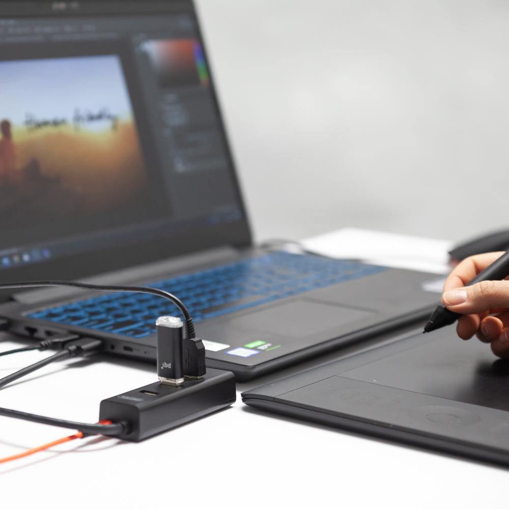 Schwarzer Unitek 4-Port-USB-Hub an einem Laptop mit einem USB-Stick, neben einem Grafiktablett mit Stift, eine Person arbeitet