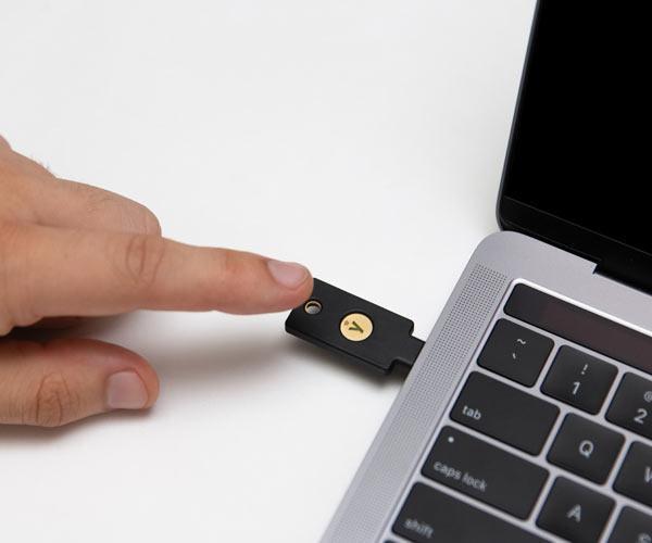 Ключ YubiKey 5C NFC, вставленный в порт USB-C MacBook, человек касается золотой кнопки для аутентификации