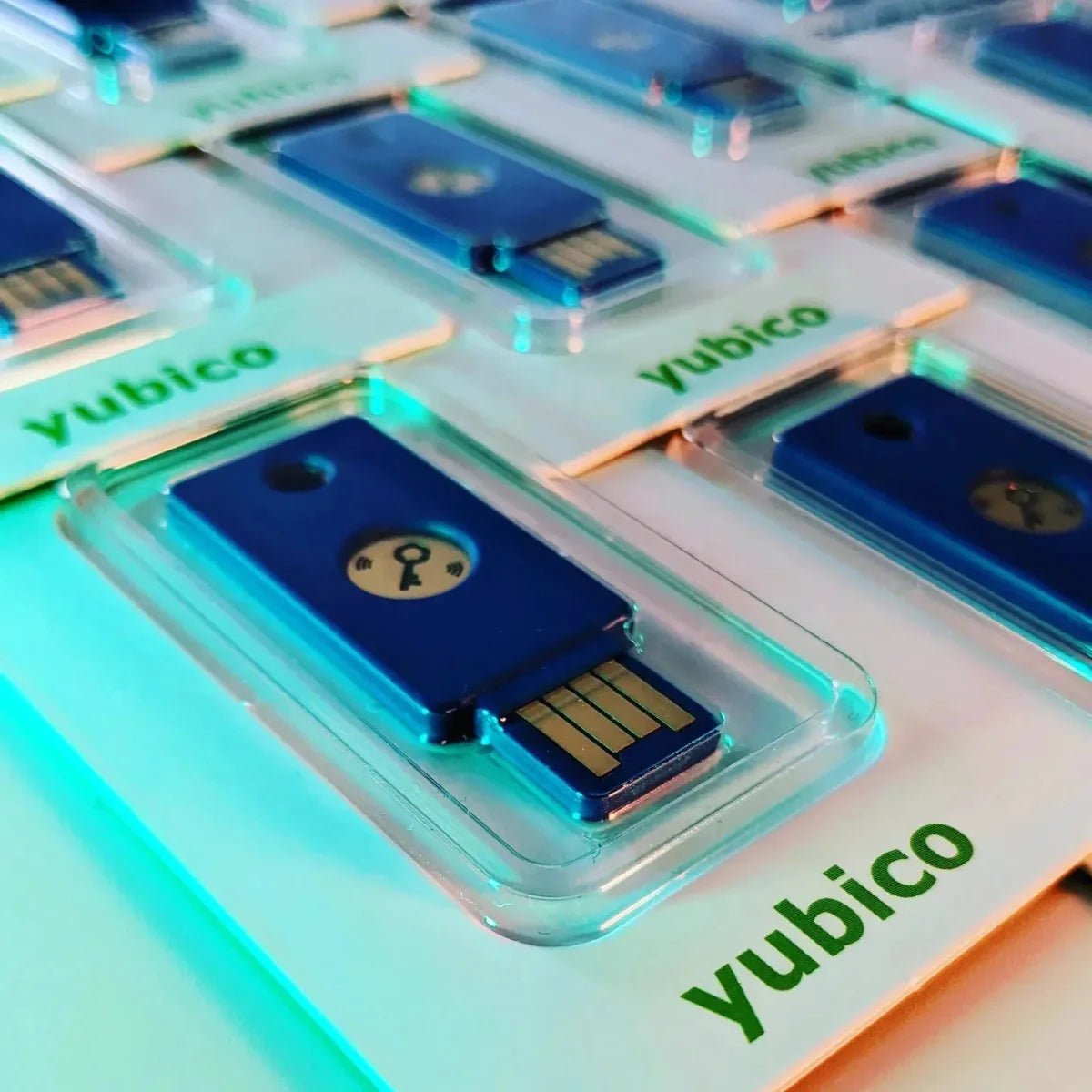 Niebieskie klucze bezpieczeństwa Yubico w blistrach ułożone rzędami, oświetlone turkusowo i pomarańczowo, zbliżenie na złocone złącze USB-A