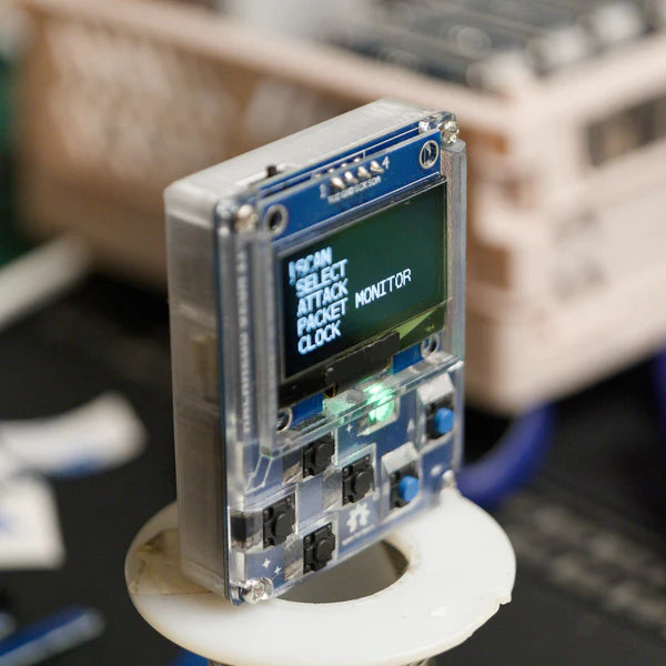 Zmontowany HackHeld Vega II+ w przezroczystej obudowie, włączony - na wyświetlaczu OLED menu: SCAN, SELECT, ATTACK, PACKET MONITOR, CLOCK, świeci zielona dioda, urządzenie na białym stojaku, rozmyte tło warsztatu