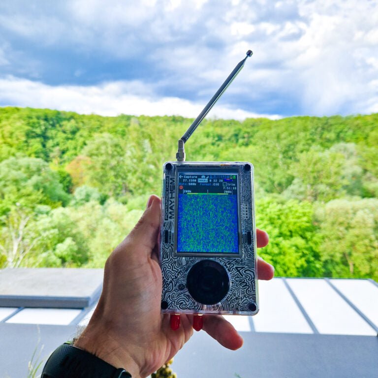 HackRF One with PortaPack H4M Mayhem held in a hand with telescopic antenna, forest in the background