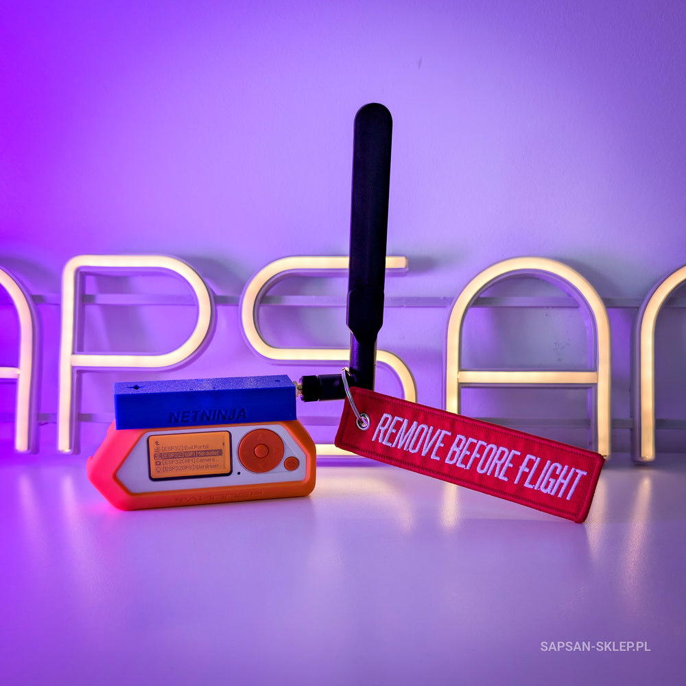 Flipper Zero with the NetNinja module and a black antenna on a white desk in front of the SAPSAN neon sign in purple light, next to a red Remove Before Flight keychain, SAPSAN-SKLEP.PL watermark