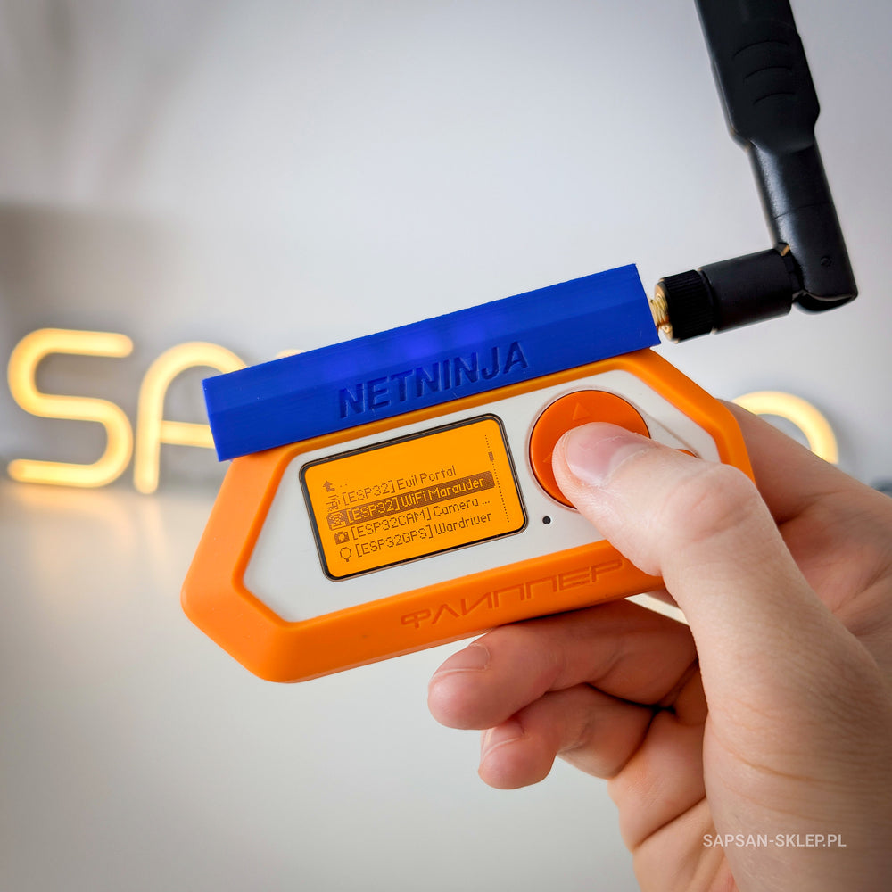 Flipper Zero in an orange case with the blue NetNinja module attached and a black antenna, held in a hand, WiFi Marauder app menu on the screen, blurred SAPSAN neon sign in the background, SAPSAN-SKLEP.PL watermark