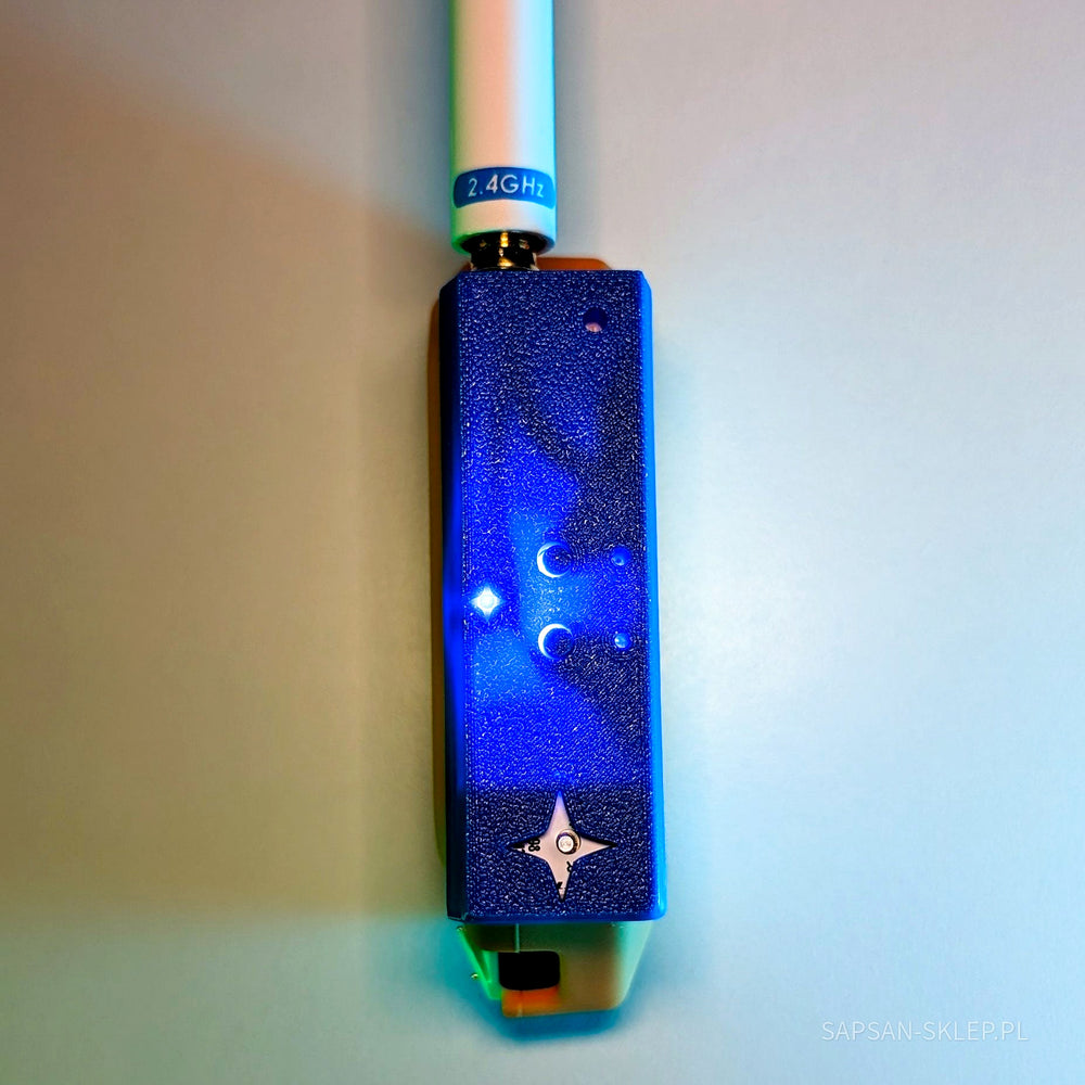 Close-up of the blue NetNinja module with a white 2.4GHz antenna screwed onto the gold SMA connector - a glowing LED on the textured enclosure and an embossed four-pointed ninja star logo, part of an orange Flipper Zero at the bottom, blue glow, SAPSAN-SKLEP.PL watermark