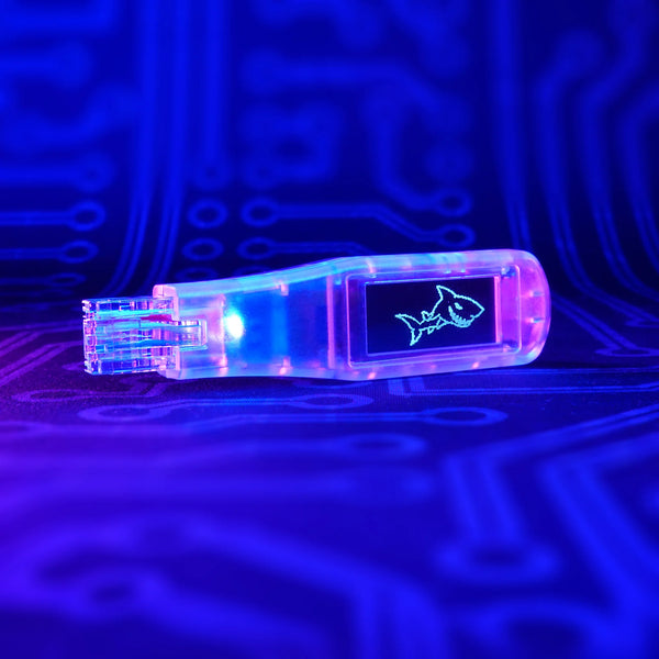 Shark Jack Display w przezroczystej obudowie ze złączem RJ45 i logo rekina na wyświetlaczu OLED, na tle płytki w świetle UV
