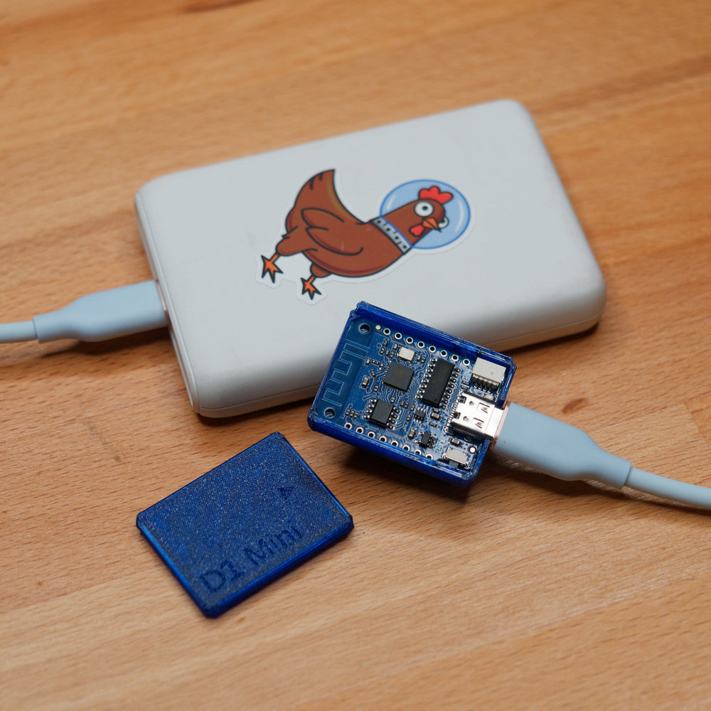 Tester WiFi D1 Mini podłączony kablem USB do power banku z dużą naklejką Spacehuhn (kogut w hełmie), obok niebieska obudowa D1 Mini, na drewnie