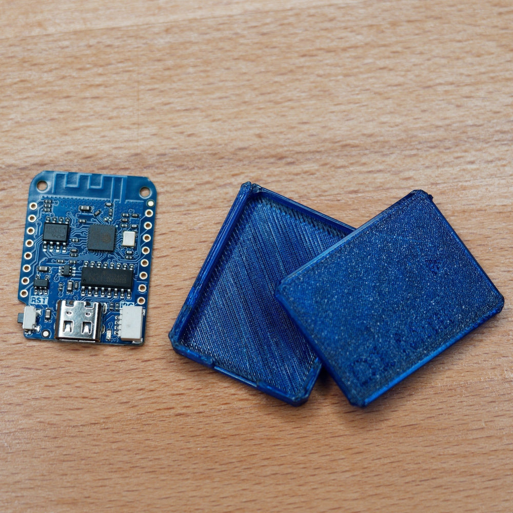 Płytka testera WiFi D1 Mini obok dwóch części niebieskiej obudowy druku 3D - na płytce układ ESP8266, ścieżka anteny WiFi, gniazdo USB i przycisk RST, na drewnie