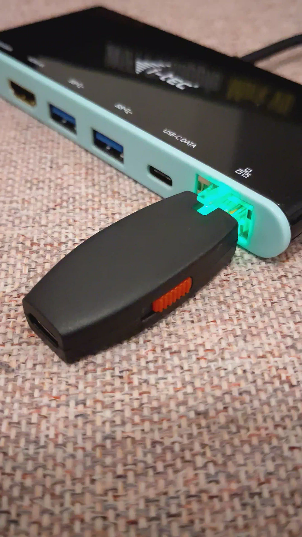 Wideo: czarny Hak5 Shark Jack z czerwonym suwakiem wpięty w port Ethernet turkusowo-czarnej stacji dokującej i-tec, port podświetlony na zielono, na beżowej tkaninie
