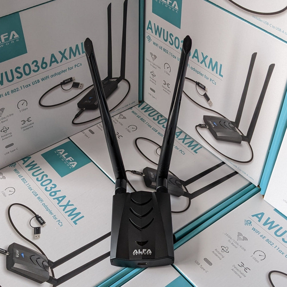 Чёрный адаптер Alfa AWUS036AXML с двумя высокими антеннами, стоящий на стопке коробок AWUS036AXML - видны логотип ALFA NETWORK и порт USB-C