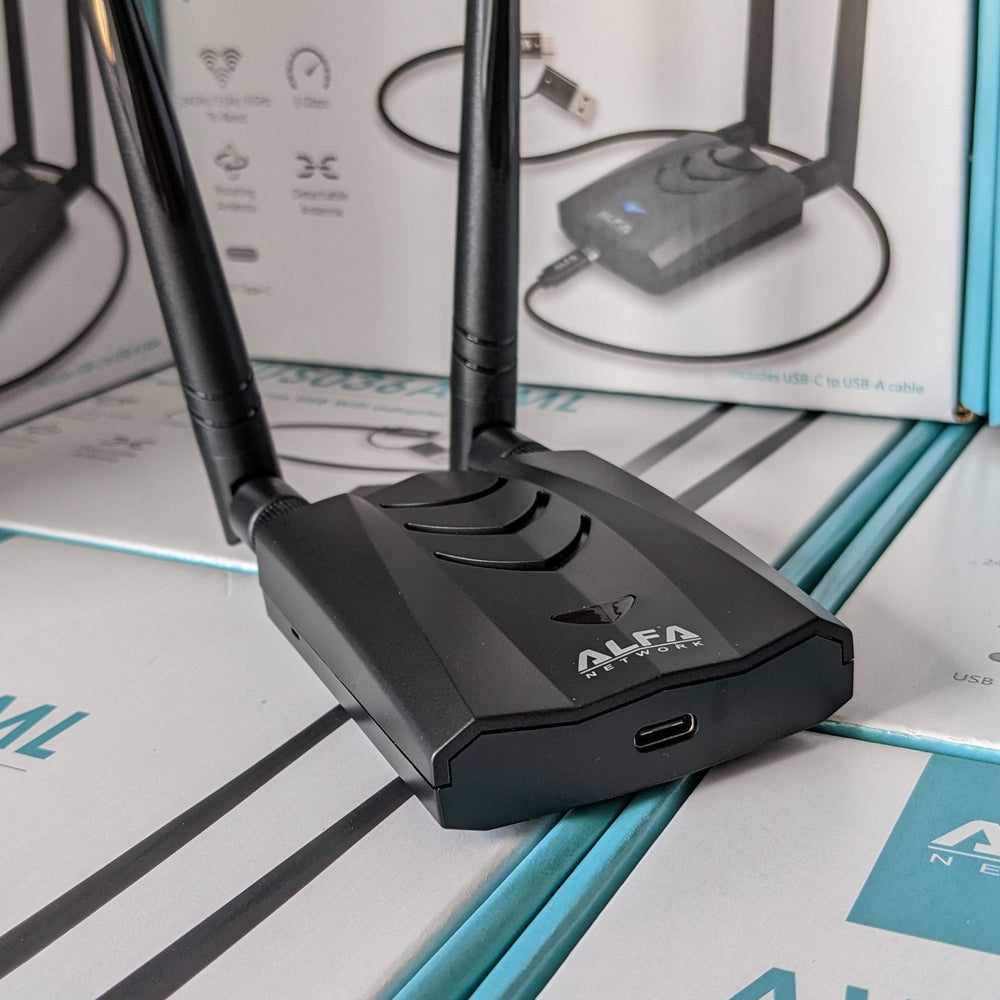 Чёрный адаптер Alfa AWUS036AXML с двумя антеннами, стоящий на упаковках продукта - видны логотип ALFA NETWORK и порт USB-C, на фоне бело-бирюзовые коробки AWUS036AXML