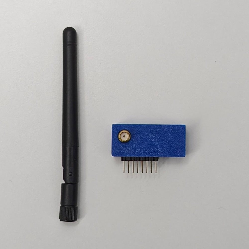 Externí modul CC1101 vedle odšroubované černé antény na světle šedém pozadí - modré pouzdro tištěné 3D se zlatým anténním konektorem SMA na horní stěně a řadou sedmi zlatých pinových konektorů vyčnívajících zespodu