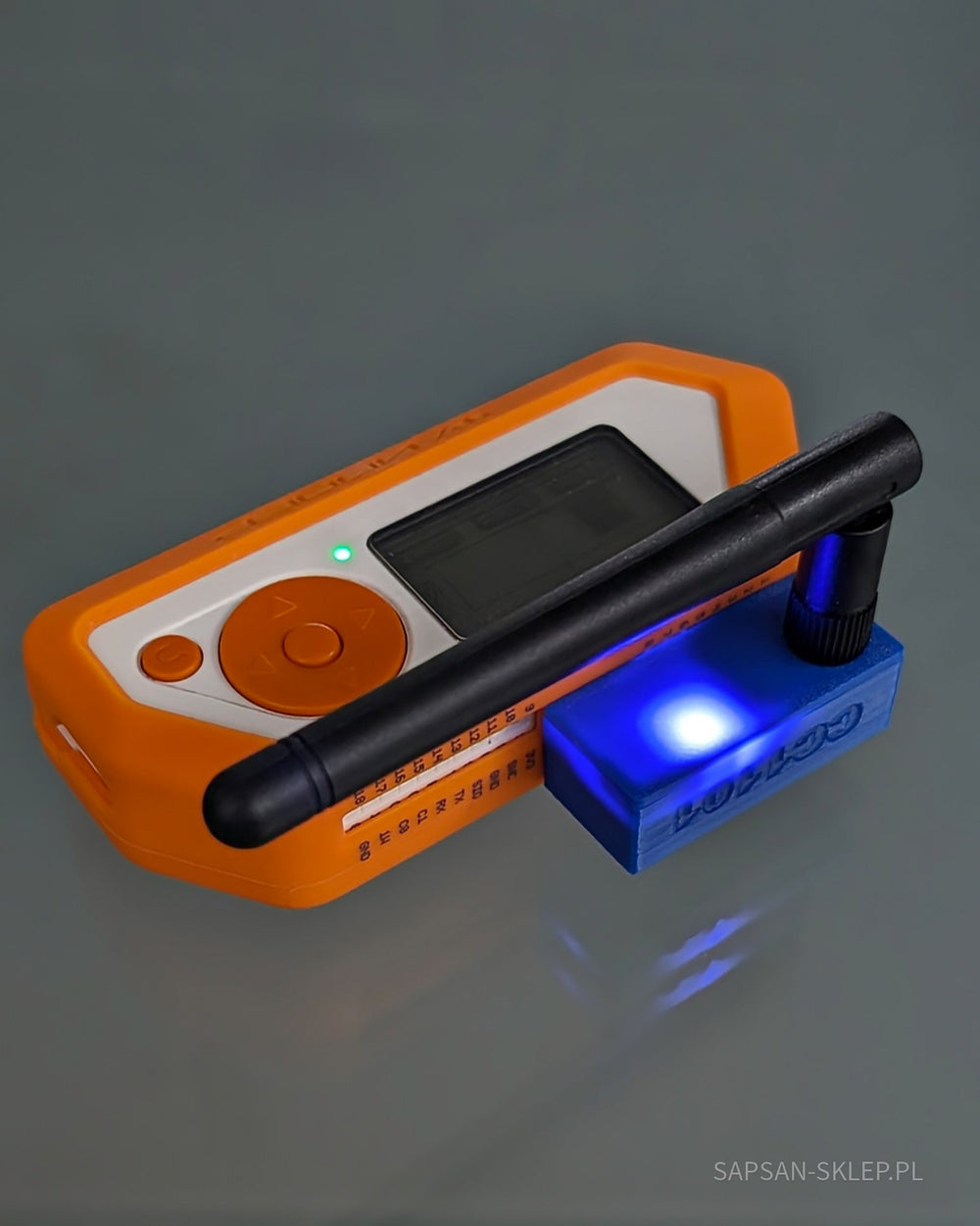 Externí modul CC1101 zapojený do pinů GPIO oranžového Flipperu Zero na šedém odrazivém pozadí - modré pouzdro tištěné 3D s vyraženým nápisem CC1101, černá anténa a svítící modrá LED, viditelné popisky pinů GPIO Flipperu a zelená LED, vodoznak SAPSAN-SKLEP.PL