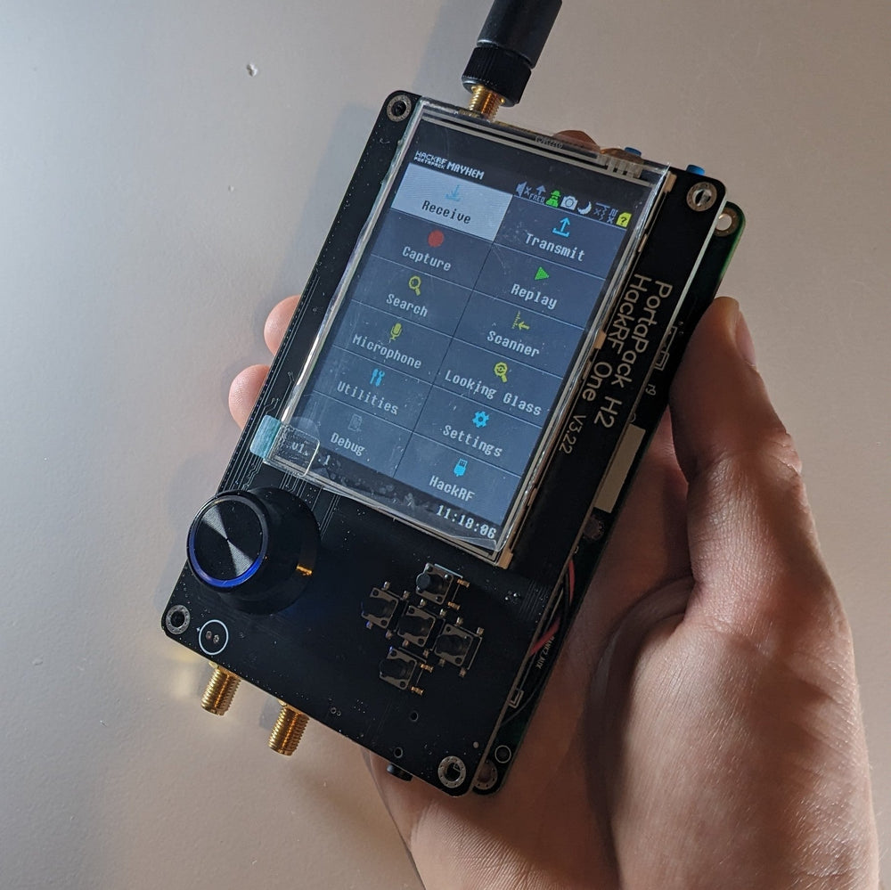 Ruka držící zařízení HackRF One s nástavbou PortaPack H2 a firmwarem Mayhem na obrazovce hlavního menu, s připojenou anténou a kolečkem