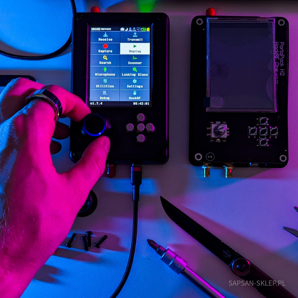 Ruka ovládající kolečko zařízení HackRF One s PortaPack H2 a firmwarem Mayhem na obrazovce menu, vedle druhá jednotka, v barevném osvětlení
