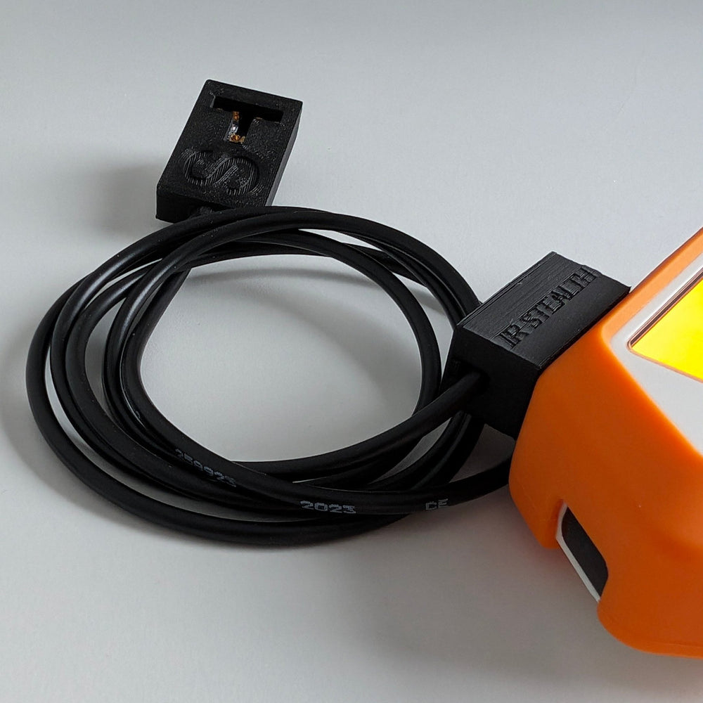 Модуль IR Stealth для Flipper Zero на светло-сером фоне - чёрный корпус, напечатанный на 3D-принтере, с тиснёной надписью IR STEALTH, свёрнутый чёрный кабель с маркировкой 2023 CE и вторая часть с инфракрасным излучателем, справа фрагмент оранжевого Flipper Zero со светящимся экраном