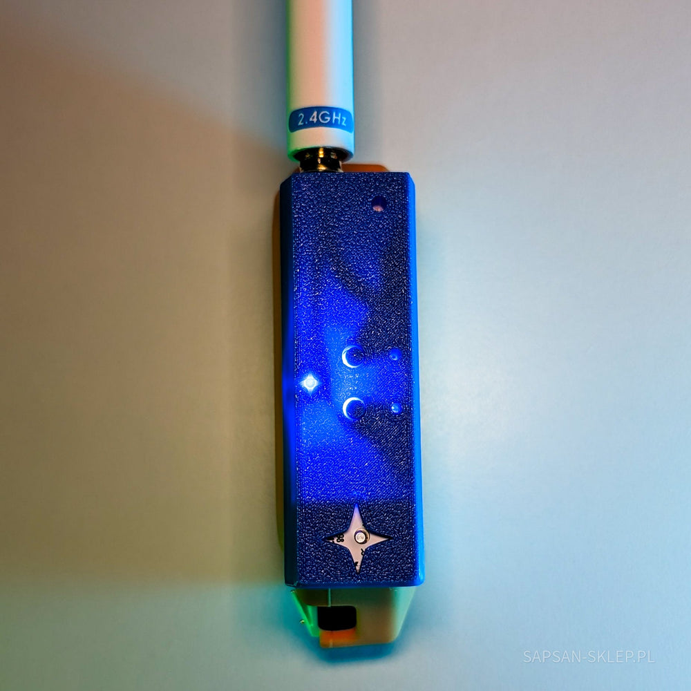 Nahaufnahme des blauen NetNinja-Moduls mit einer weißen 2.4GHz-Antenne, die über einen goldenen SMA-Anschluss aufgeschraubt ist - eine leuchtende LED und ein eingeprägtes vierzackiges Ninjastern-Logo auf dem texturierten Gehäuse, unten ein Ausschnitt eines orangefarbenen Flipper Zero, ein blaues Leuchten, das SAPSAN-SKLEP.PL-Wasserzeichen