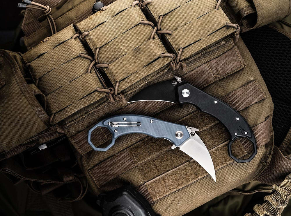 Zwei geöffnete Böker Plus HEL Karambit Messer - schwarz mit schwarzer Klinge und blaugrau mit Satin-Klinge - überkreuzt auf einem sandfarbenen Plattenträger mit Magazintaschen und MOLLE-System