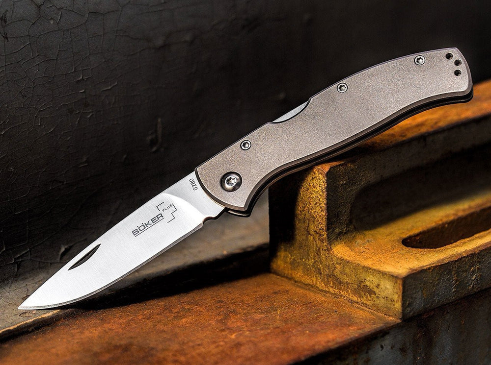 Lifestyle photo of the open Böker Plus Titan Drop knife with a satin drop point blade and a grey titanium handle on a rusty metal background