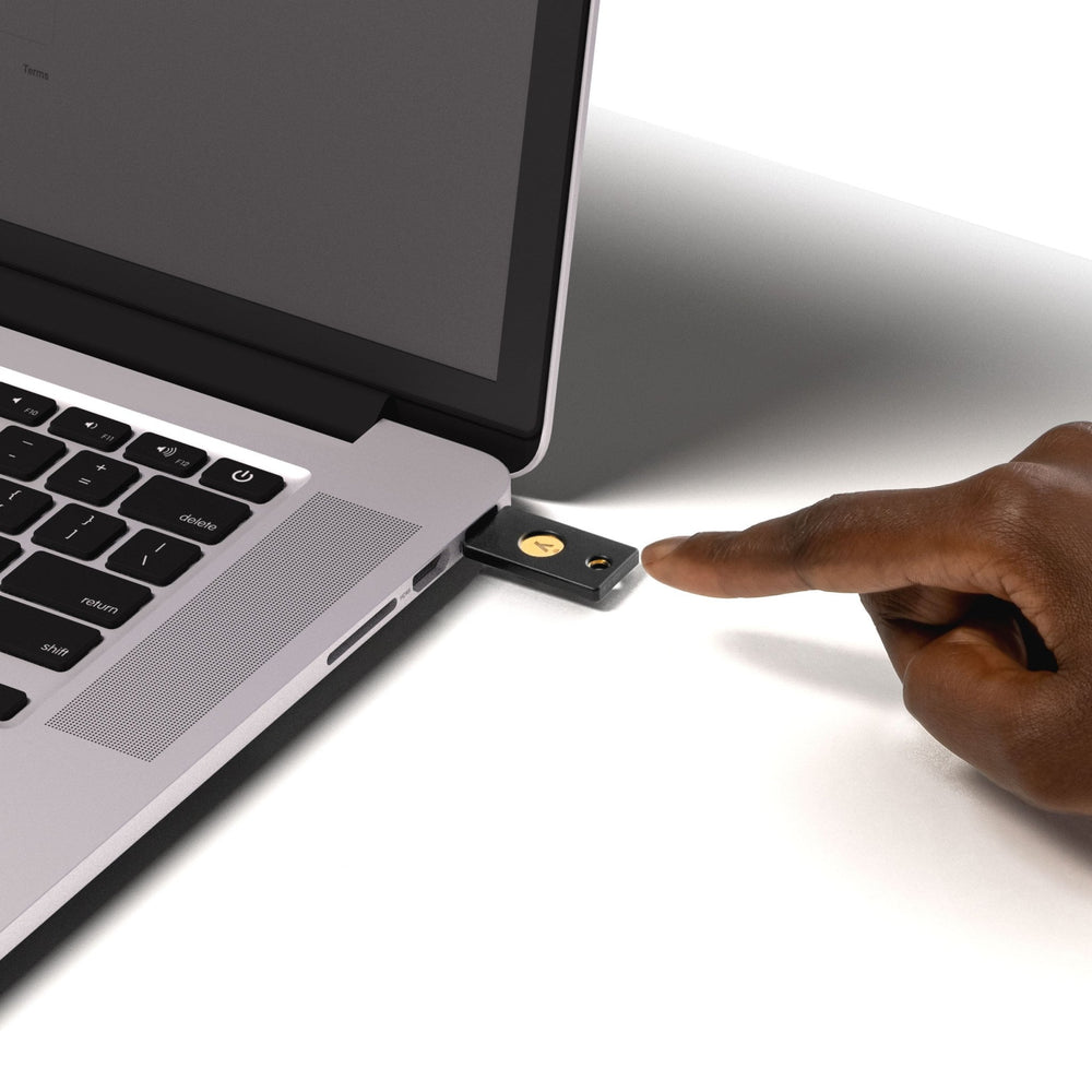 Klíč Yubico Security Key NFC zapojený do portu USB-A MacBooku, osoba se dotýká zlatého tlačítka pro ověření