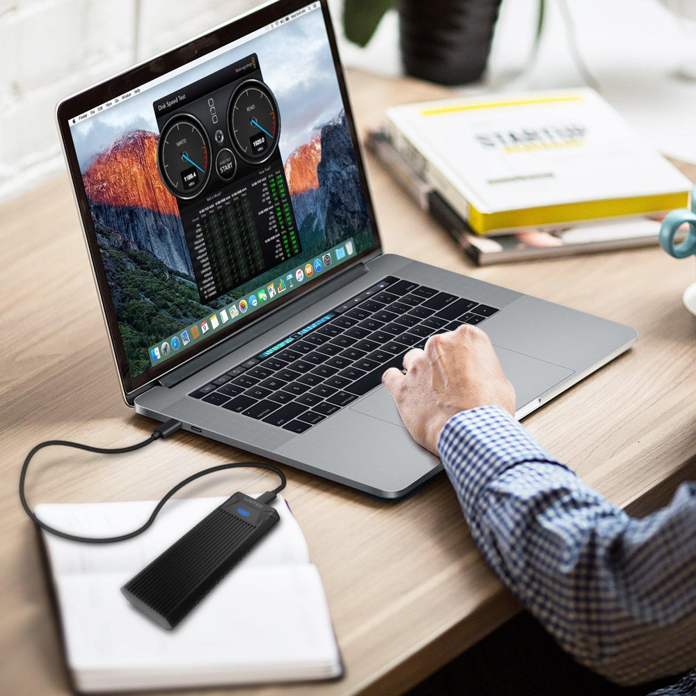 Unitek M.2 Laufwerksgehäuse an einem MacBook auf dem Schreibtisch, eine Person arbeitet, ein Laufwerks-Geschwindigkeitstest auf dem Bildschirm