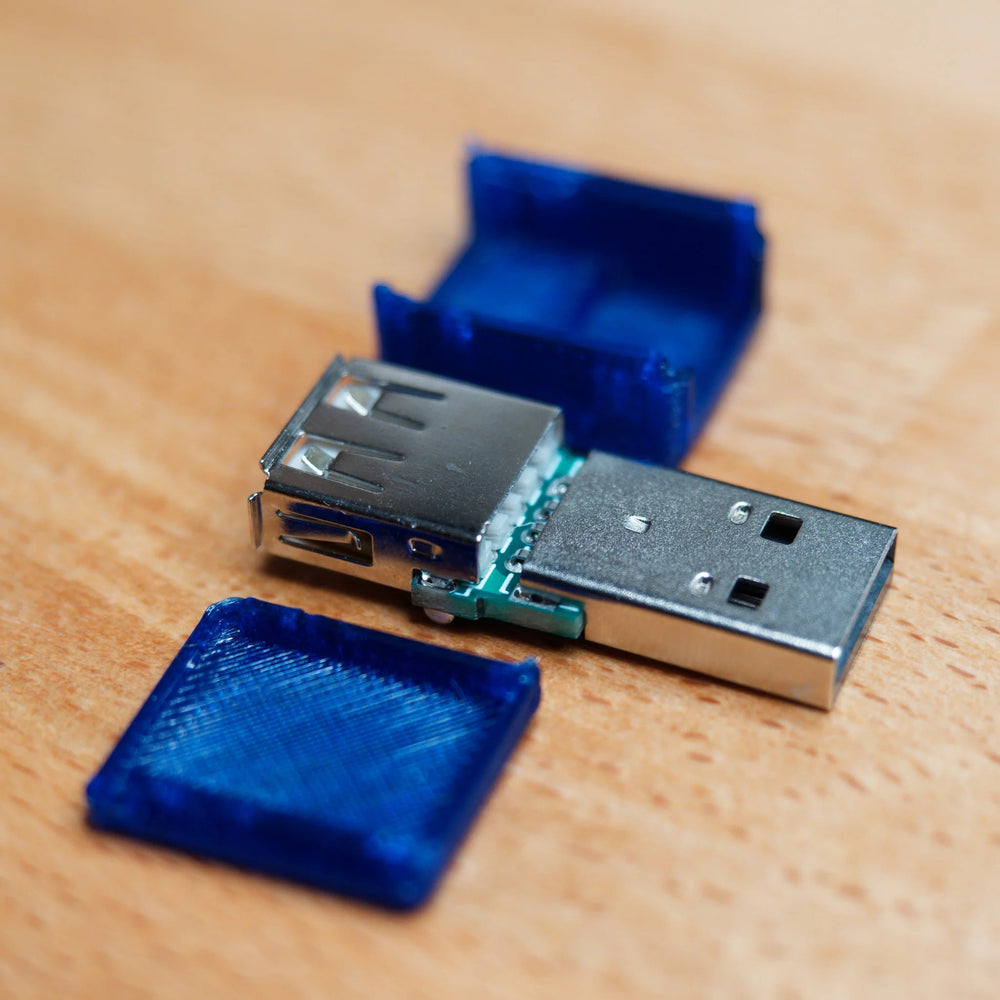 USB blokátor dat rozložený - viditelná zelená deska spojující konektor a zásuvku USB-A (jen napájecí piny), vedle dvě poloviny modrého pouzdra, na dřevě