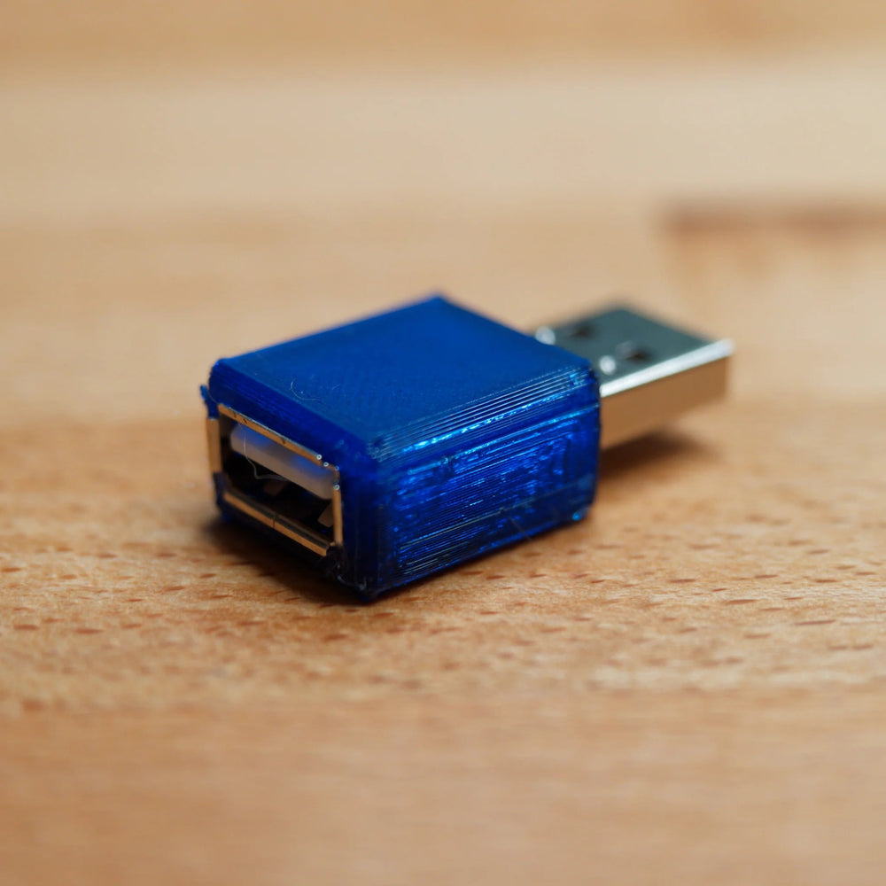 USB blokátor dat - viditelné oba konce: konektor USB-A a zásuvka USB-A v průhledném modrém pouzdře z 3D tisku, na dřevě