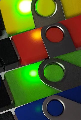 Zbliżenie kolorowych obudów USB Rubber Ducky z prześwitującym światłem LED - ułożone pod kątem obudowy żółta, czerwona, zielona i niebieska ze stalowymi obrotowymi osłonami, każda podświetlona wewnętrzną diodą, na ciemnym tle