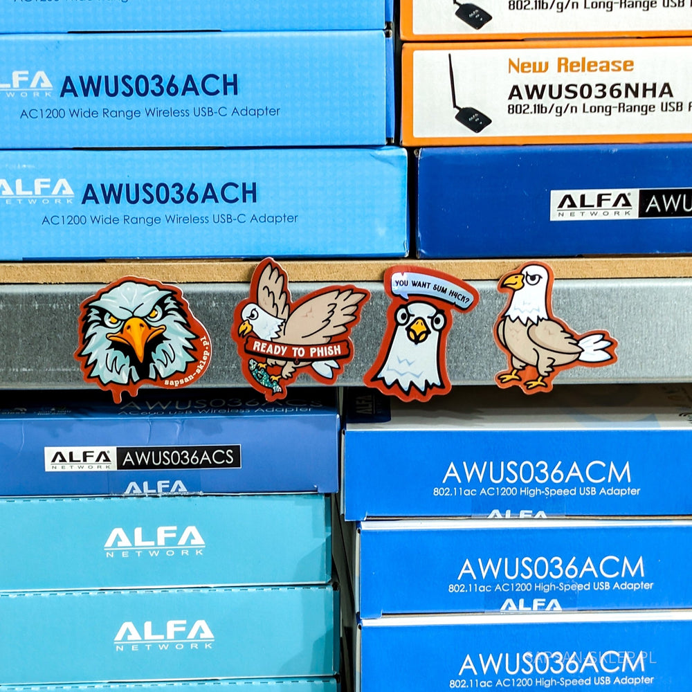 Four SAPSAN stickers standing on the edge of a shelf between stacked ALFA Network WiFi adapter boxes (AWUS036ACH AC1200 USB-C, AWUS036NHA, AWUS036ACS, AWUS036ACM) - stickers with the eagle mascots: an angry eagle head with the sapsan-sklep.pl text, an eagle with a READY TO PHISH banner and a fish, an eagle with a YOU WANT SUM H4CK speech bubble and a standing eagle, the SAPSAN-SKLEP.PL watermark