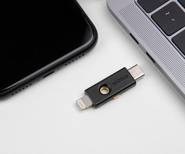 Schwarzer YubiKey 5Ci mit USB-C- und Lightning-Steckern zwischen einem iPhone und einem MacBook, zeigt die Kompatibilität mit beiden Geräten