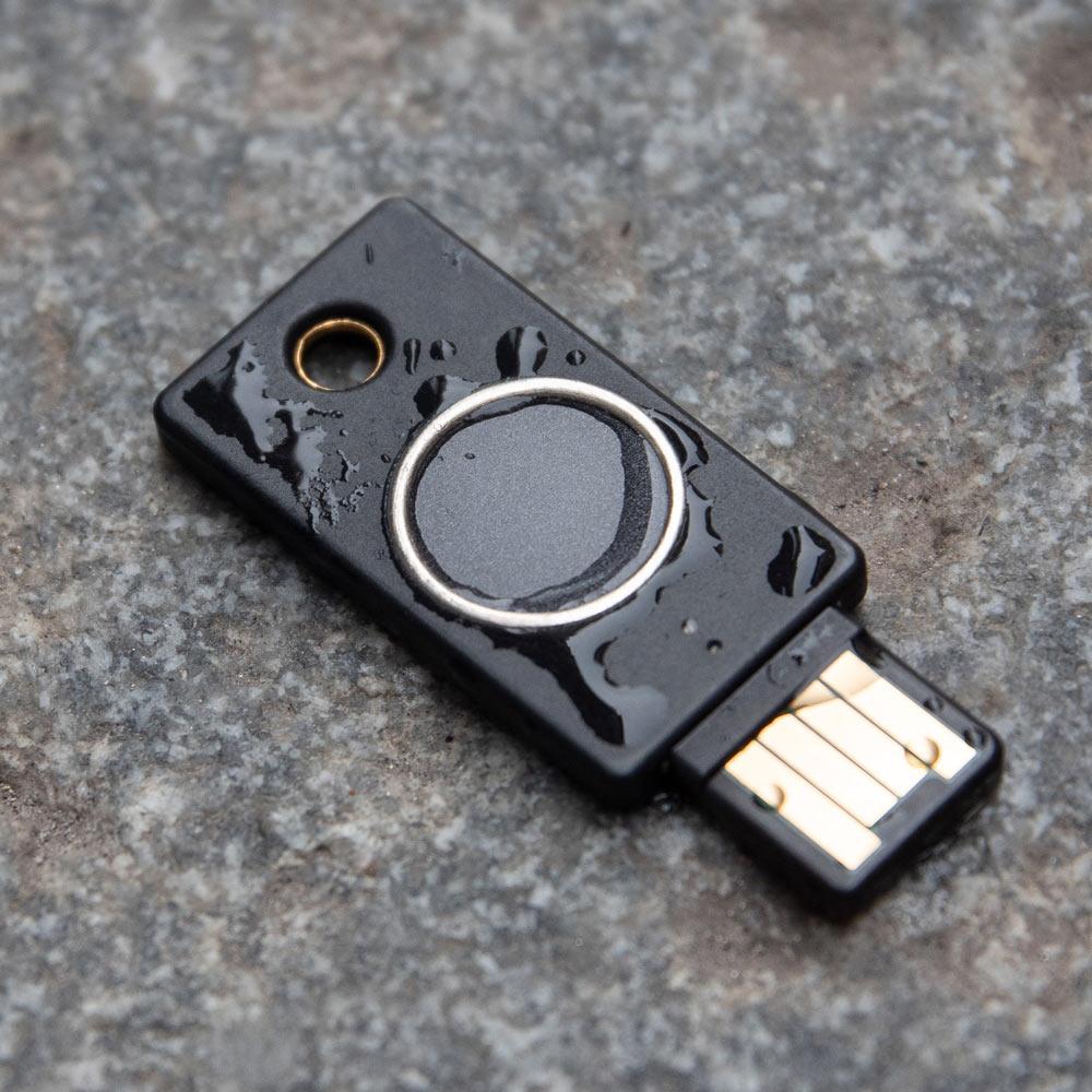 Schwarzer YubiKey Bio Schlüssel mit biometrischem Fingerabdruckleser und USB-A-Stecker, mit Wassertropfen bedeckt auf einer Steinoberfläche