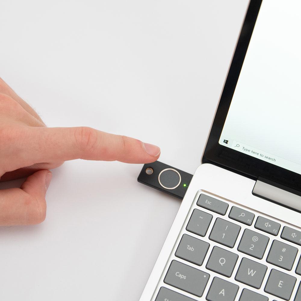 Nutzungsszene - schwarzer YubiKey Bio Schlüssel im USB-A-Anschluss eines Laptops, ein Finger berührt den biometrischen Fingerabdruckleser mit einer leuchtenden grünen LED