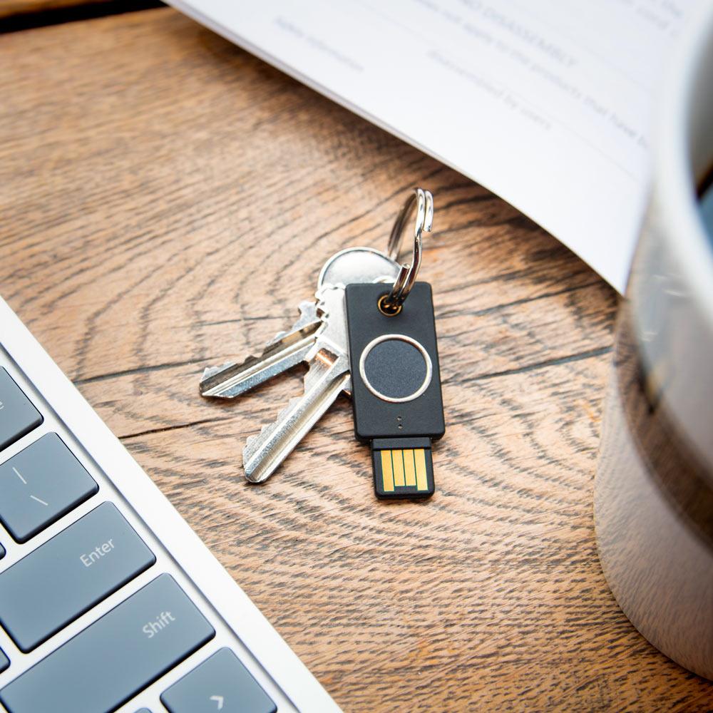 Schwarzer YubiKey Bio Schlüssel an einem Schlüsselring auf einem Holzschreibtisch neben einem Laptop, Dokumenten und einer Tasse