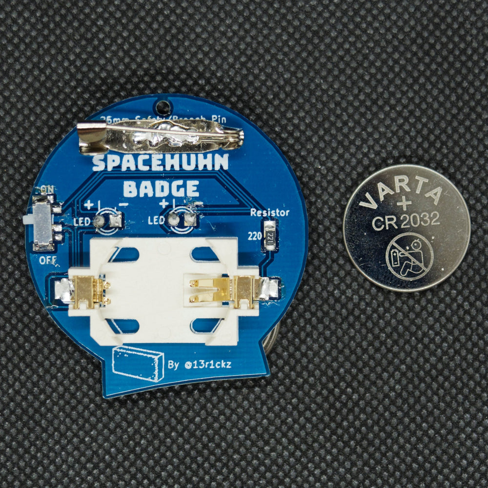 Die Rückseite eines Spacehuhn-Abzeichens - eine blaue Platine mit der Aufschrift SPACEHUHN BADGE, einer Sicherheitsnadel, einem ON/OFF-Schalter, zwei LED-Plätzen, einem 220-Widerstand und einem Knopfzellenhalter, daneben eine VARTA-CR2032-Knopfzelle
