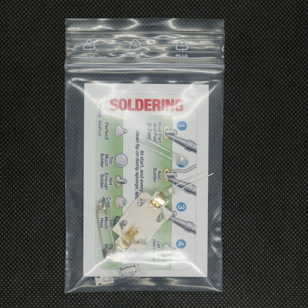 Die Spacehuhn-Bauteile in einem transparenten Zip-Beutel - eine SOLDERING-Anleitungskarte, LEDs, ein Batteriehalter, ein Schalter und eine Sicherheitsnadel