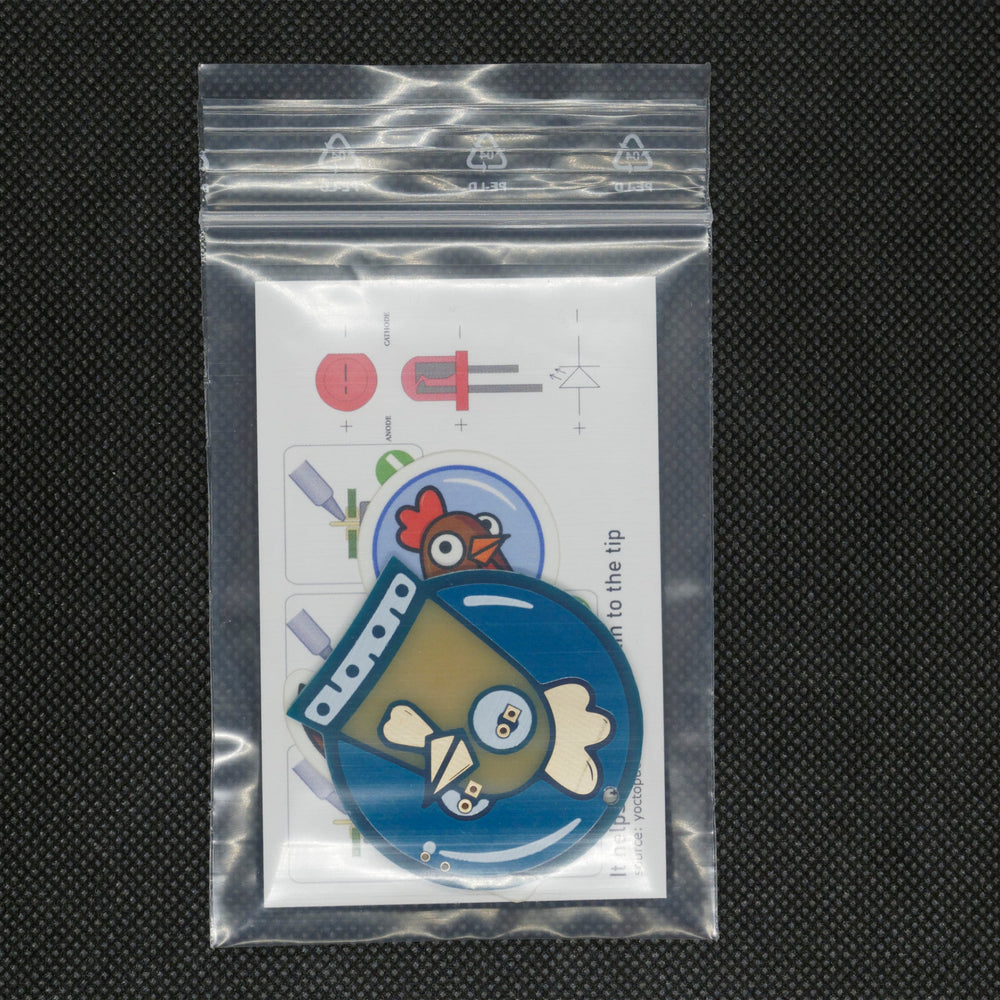 Ein komplettes Spacehuhn-Set im Zip-Beutel - die unmontierte Hahn-Abzeichenplatine, ein Aufkleber und eine Karte mit LED-Polarität (Kathode/Anode) und Löttipps