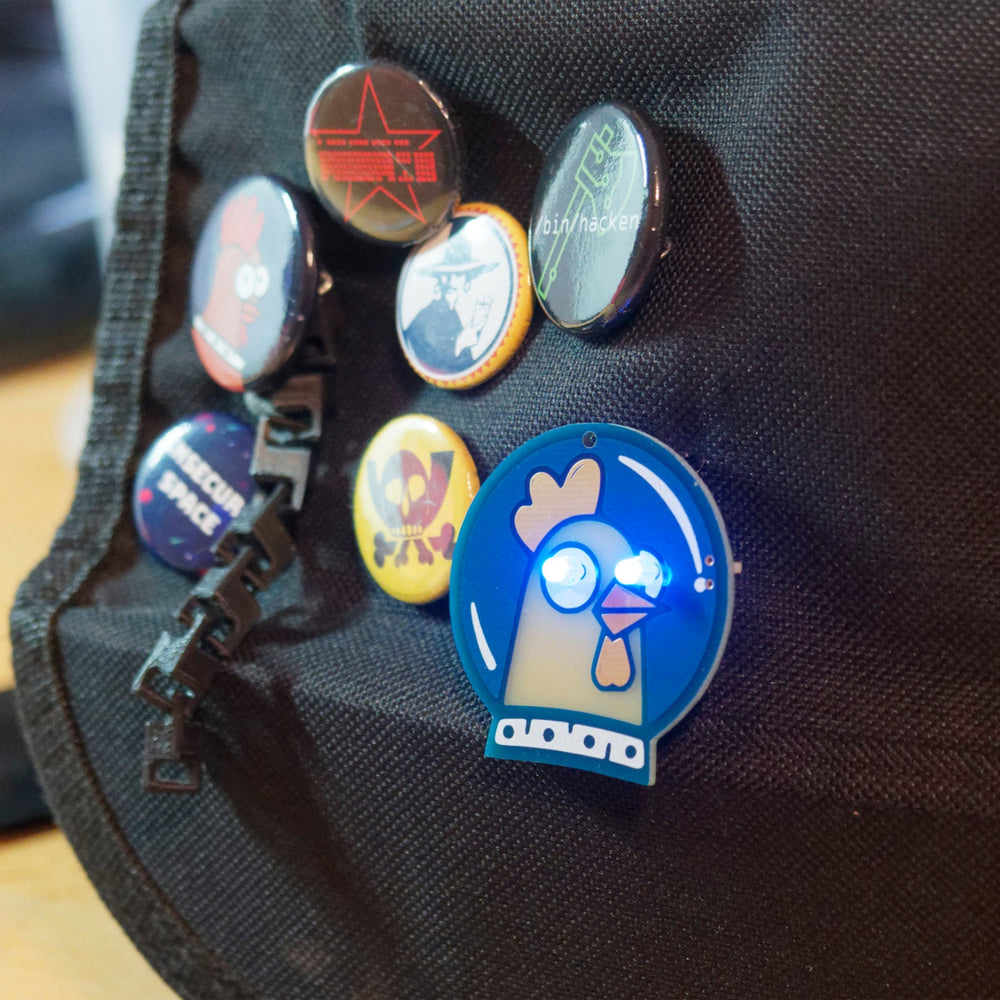 Ein Spacehuhn-Abzeichen mit blau leuchtenden Augen an einem schwarzen Rucksack neben anderen Hacker-Ansteckern (ein roter HACK-Stern, /bin/hacken, ein Totenkopf, INSECUR SPACE)